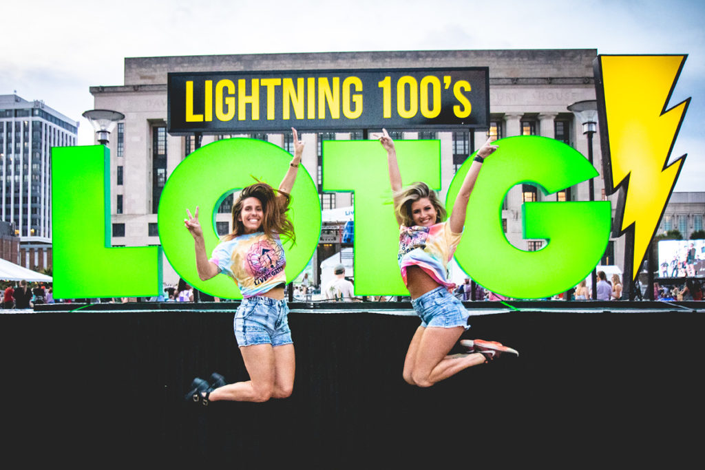 Two models jumping in Live On The Green tees in front of the LED sign